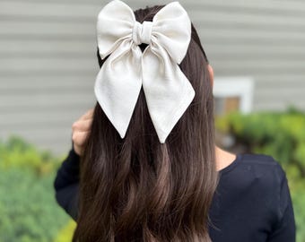 May include: A large, off-white hair bow is centered on a person's dark brown hair. The bow has a textured appearance and is tied in the center. The person is wearing a black top. The background is blurred, with green foliage and a gray wall visible.