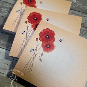 May include: Three square wooden notepads with a floral design. Each notepad features a red poppy flower with a dark center, accompanied by a few small bees. The notepads have a light brown background and a grid pattern.