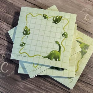 May include: A stack of square sticky notes with a light green grid pattern. Each note features a green dinosaur illustration with a long neck and a leafy border. The notes are stacked on a wooden surface.
