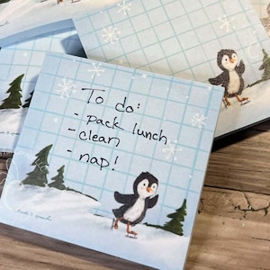 Puede incluir: Notas adhesivas cuadradas de color azul claro con una escena invernal con un pingüino patinando sobre hielo. Las notas tienen un patrón de cuadrícula y copos de nieve. Una nota tiene una lista de tareas manuscrita.