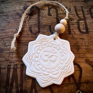 May include: A white ceramic ornament with a mandala design and the Om symbol in the center. It has a natural twine hanger with two wooden beads. The ornament is set against a rustic wooden background.