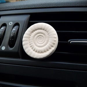 May include: A circular, cream-coloured car air freshener featuring a spiral fossil pattern. The air freshener is positioned on a black car vent, offering a decorative and practical addition to the vehicle's interior.