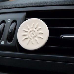 Op de afbeelding: Een ronde, gebroken witte auto luchtverfrisser met een zonontwerp. De zon heeft een spiraalvormig midden en stralen. De verfrisser is bevestigd aan een zwart ventilatierooster, met de bedieningselementen zichtbaar.