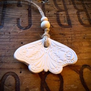 Könnte beinhalten: Weißes, schmetterlingsförmiges Ornament mit aufwendigen geprägten Details, aufgehängt an einer natürlichen Schnur und Holzperlen. Das Ornament hat ein detailliertes Flügelmuster und befindet sich vor einem rustikalen Holzgrund.