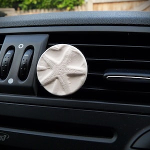 May include: A circular, off-white car air freshener featuring a starfish design. It's attached to a black car vent. The vent has adjustable airflow controls. The air freshener is approximately 7 cm in diameter.
