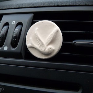 Op de afbeelding: Een ronde, crèmekleurige auto-luchtverfrisser met een gedetailleerd reliëf van een vogel in vlucht. De luchtverfrisser is geplaatst op een zwarte ventilatieopening in de auto, met de lamellen en knoppen op de achtergrond.