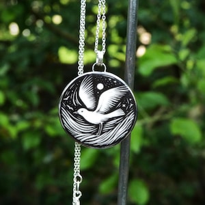 May include: A silver necklace with a round pendant featuring a white bird in flight against a black background. The design includes stars, a moon, and stylized waves. The chain is delicate and hangs against a blurred green backdrop.