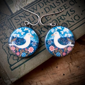 May include: Round earrings with a white seagull design, set against a blue background with pink and blue flowers. The earrings have silver-colored hooks and are displayed on a vintage book. The earrings are a unique accessory.