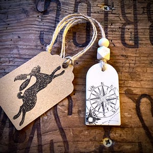 Könnte beinhalten: Zwei Anhänger: Einer mit einer springenden Hasenillustration, der andere mit einem Kompassrosen-Design. Beide sind mit Schnur und Holzperlen befestigt. Auf einer rustikalen Holzoberfläche.