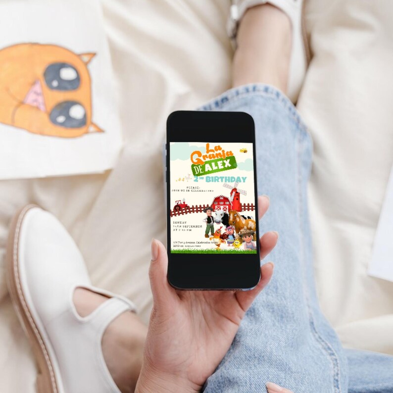 May include: A smartphone displays a colorful birthday invitation with the text "La Granja de Alex 4th Birthday." The invitation features cartoon farm animals, a red barn, and a tractor. The phone is held in a person's hand, with a drawing and white shoe visible.