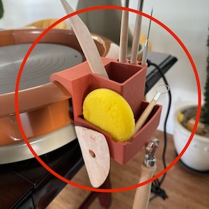 May include: A terracotta-coloured pottery tool organiser attached to a pottery wheel. The organiser holds various tools, including wooden ribs, metal tools, and a yellow sponge. A wooden tool and a metal tool with a wooden handle are also visible.