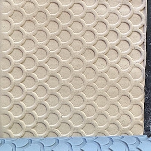 May include: A beige clay slab with a repeating scale pattern is shown, with a blue roller below. The detailed texture suggests a tool for creating patterned surfaces, possibly for pottery or crafts. The scale pattern is a repeating design.