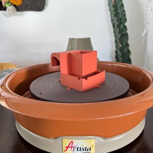 May include: An orange and gray pottery wheel with a dark brown rotating surface. A small orange structure sits on the wheel. The wheel has the word "Artista" printed on it.