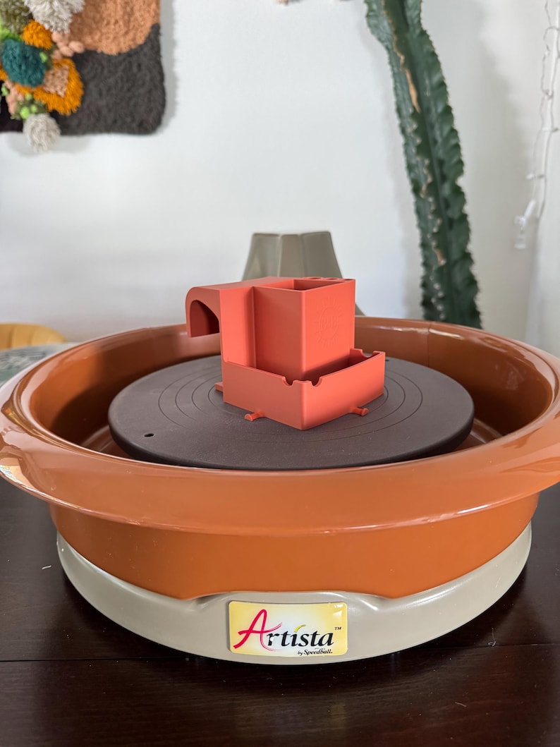 May include: An orange and gray pottery wheel with a dark brown rotating surface. A small orange structure sits on the wheel. The wheel has the word "Artista" printed on it.