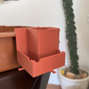 May include: A terracotta-colored organizer with a rectangular shape and multiple compartments. The organizer has a small tray and the letters "B&B" inside a sun design. It is designed to attach to a larger container.