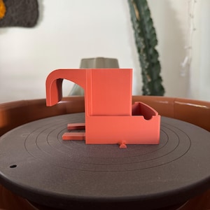 May include: A coral-colored, 3D-printed object with a curved handle and a rectangular base. The object sits on a dark gray, circular surface with concentric grooves, possibly a record player. The overall design is modern and minimalist.