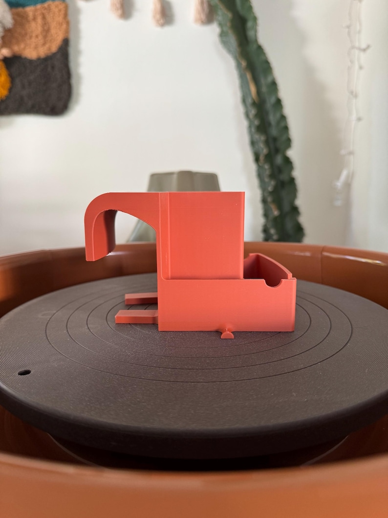 May include: A coral-colored, 3D-printed object with a curved handle and a rectangular base. The object sits on a dark gray, circular surface with concentric grooves, possibly a record player. The overall design is modern and minimalist.