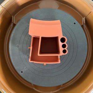 May include: An orange pottery wheel tool holder with three circular holes and a rectangular compartment. The tool holder sits on a gray surface with concentric circles, inside a brown pottery wheel.