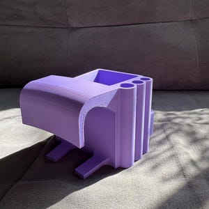 May include: A 3D-printed, lavender-colored desk organizer. The organizer has a rectangular shape with a curved top and several cylindrical compartments. It features a small shelf and two legs for stability. The object is made of plastic.