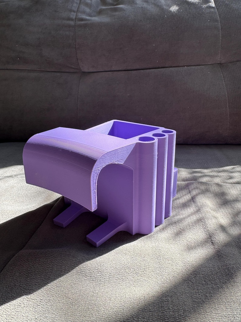 May include: A 3D-printed, lavender-colored desk organizer. The organizer has a rectangular shape with a curved top and several cylindrical compartments. It features a small shelf and two legs for stability. The object is made of plastic.