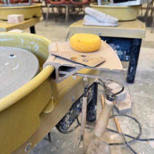 May include: A pottery wheel with attached tools and a sponge. The wheel is a light yellow colour, with a grey work surface. A wooden tool and a sponge sit on a shelf attached to the wheel. Two additional tools hang below the shelf.
