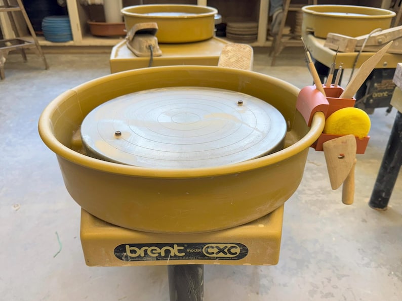 May include: A yellow pottery wheel with a rotating metal surface. The wheel has a built-in splash pan and a tool holder with various pottery tools. The base of the wheel has the brand name "brent" and model "cxc".