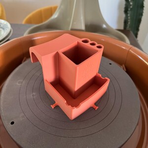 May include: A coral-colored pottery tool holder with a square container, a small tray, and several circular holes. The tool holder is sitting on a pottery wheel. The word "Tori" is visible on the side of the tool holder.