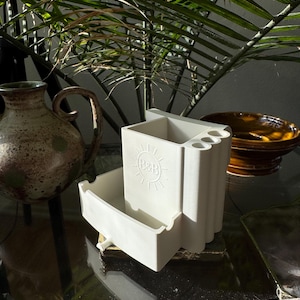 May include: A white ceramic desk organizer with a drawer and compartments for pens and pencils. The organizer has a sun logo and is sitting on a glass surface. A ceramic vase and a bowl are in the background.