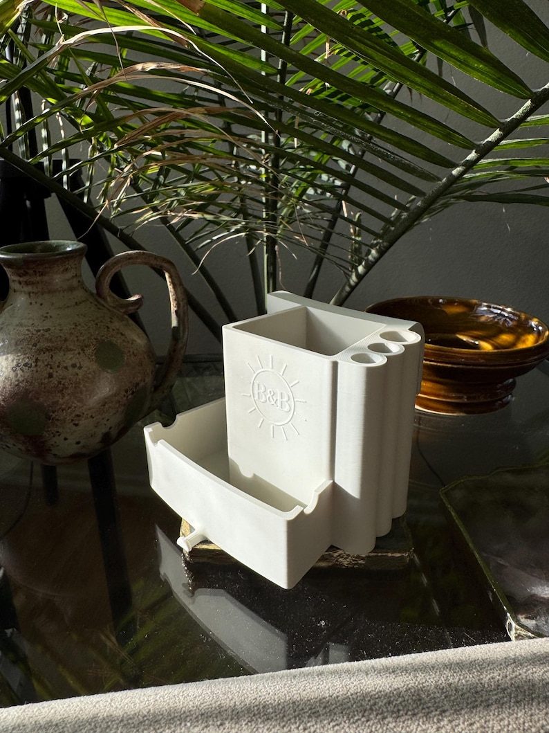 May include: A white ceramic desk organizer with a drawer and compartments for pens and pencils. The organizer has a sun logo and is sitting on a glass surface. A ceramic vase and a bowl are in the background.