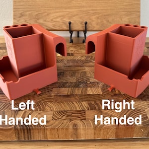 May include: Two terracotta-colored organizers, labeled "Left Handed" and "Right Handed", sit on a wooden cutting board. Each organizer has a rectangular compartment, a curved handle, and small pegs. The right-handed organizer has a floral design.