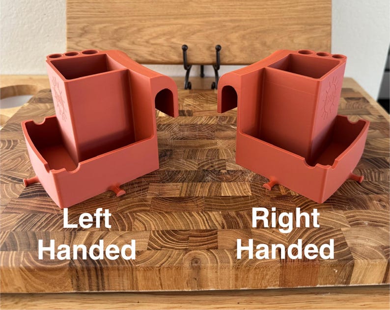 May include: Two terracotta-colored organizers, labeled "Left Handed" and "Right Handed", sit on a wooden cutting board. Each organizer has a rectangular compartment, a curved handle, and small pegs. The right-handed organizer has a floral design.