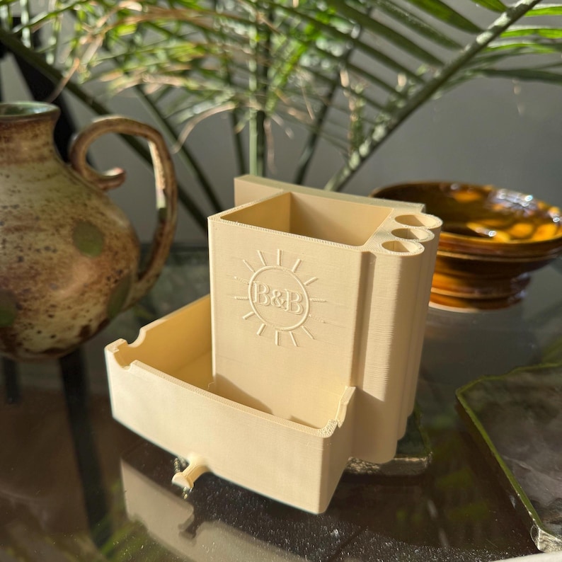 May include: A beige, 3D-printed desk organizer with a rectangular shape and a drawer. The organizer has a sun and "B&B" logo. The design includes compartments for pens and other small items. The organizer is made of plastic.
