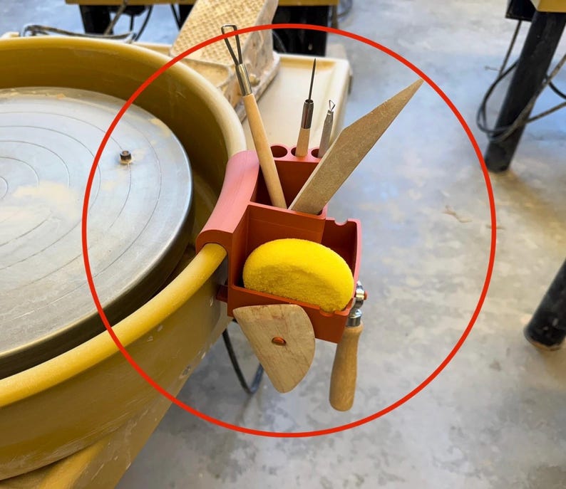 May include: A red and pink pottery tool holder attached to a pottery wheel. The holder contains various tools, including a wooden rib, a sponge, and several metal tools with wooden handles. The pottery wheel is beige and has a yellow hose.