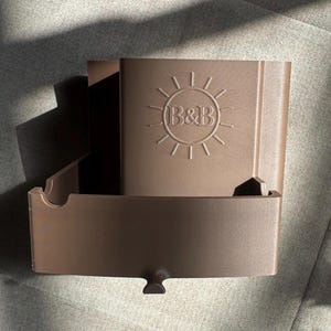 May include: A brown plastic organizer with a sun logo and the letters "B&B" embossed on the front. The organizer has a rectangular shape with an open compartment and a small, curved handle at the bottom.