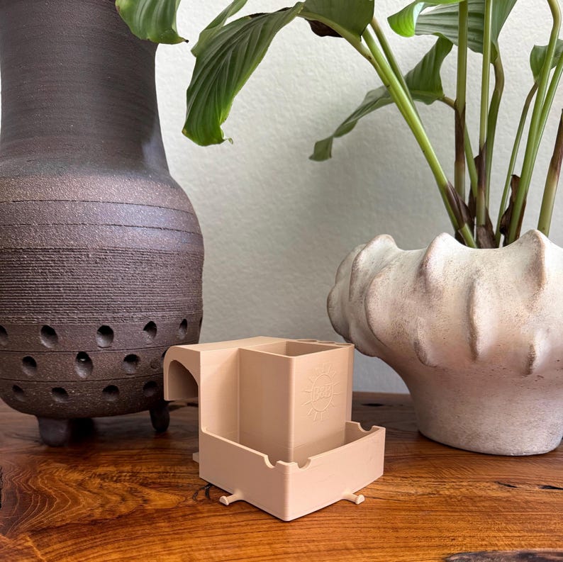 May include: A beige, geometric planter with a sun design, featuring a rectangular base and a raised square section. The planter is on a wooden surface, with a dark brown vase and a white, textured pot with a plant in the background.