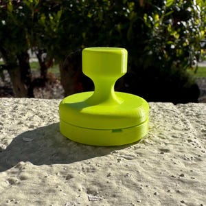 May include: A bright neon green stamp with a round base and a handle. The stamp is sitting on a textured surface, with a blurred green background.