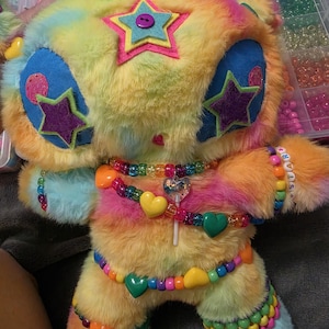 May include: A plush toy with rainbow-colored faux fur, featuring large blue eye patches with purple star appliqués. A felt star with a purple button sits on the forehead. The toy is adorned with colorful plastic bead necklaces and bracelets, including heart-shaped beads and a small heart charm.