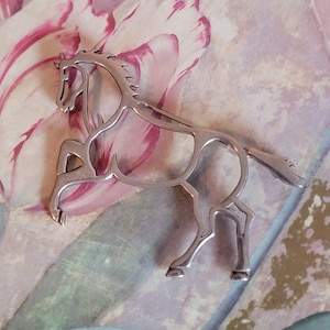 May include: A silver-colored horse brooch. The brooch depicts a stylized horse in a running pose, with an open design. The horse's mane and tail are detailed. The brooch is likely made of metal and is a piece of jewelry.