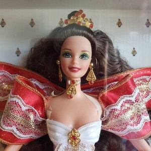 May include: A brunette Barbie doll wearing a red and white gown with gold accents and a crown. She has green eyes and red lipstick.