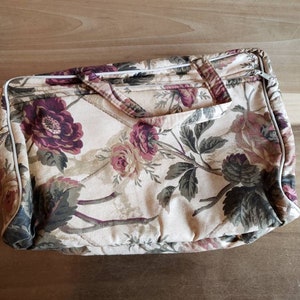 May include: A beige floral patterned zippered travel bag with a white trim. The bag has a large floral print with pink, red, and green flowers.