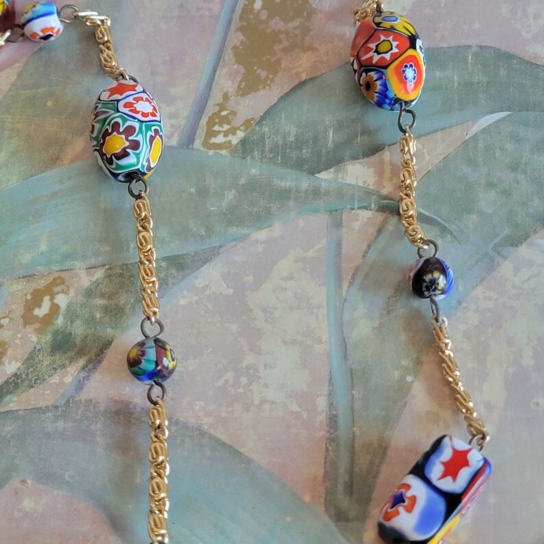 May include: A gold chain necklace with colorful glass beads. The beads are round and oval shaped and feature a variety of floral and geometric patterns.