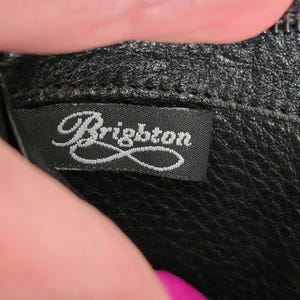 May include: A black leather accessory featuring a textured surface and a black fabric interior label with the word Brighton embroidered in white script lettering. The label is securely stitched into the seam of the black leather material.