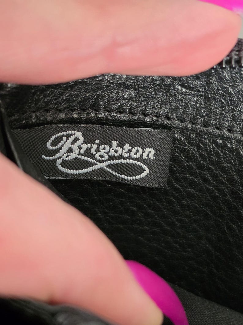 May include: A black leather accessory featuring a textured surface and a black fabric interior label with the word Brighton embroidered in white script lettering. The label is securely stitched into the seam of the black leather material.