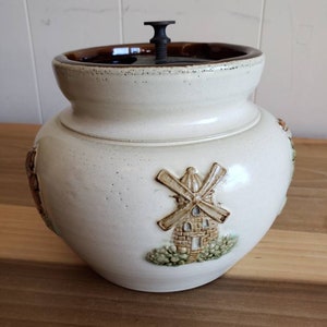 May include: Cream-colored ceramic jar with a brown rim and lid, featuring a windmill design. The jar has a bulbous shape and a dark metal handle on the lid. The windmill is embossed on the side.