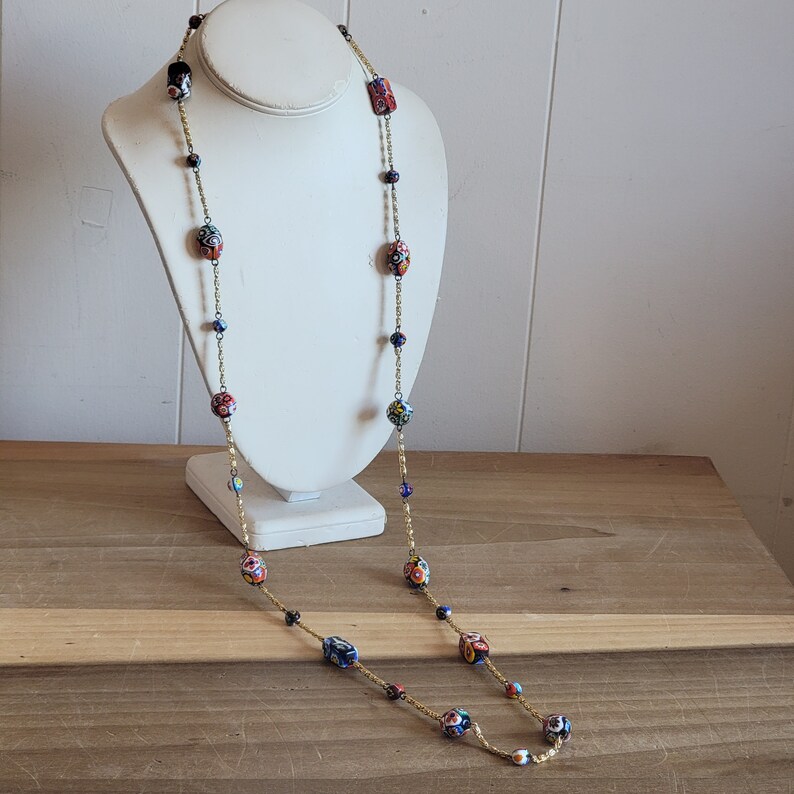 May include: A long gold chain necklace with colorful glass beads. The beads are a mix of red, blue, and yellow with intricate patterns. The necklace is displayed on a white mannequin.