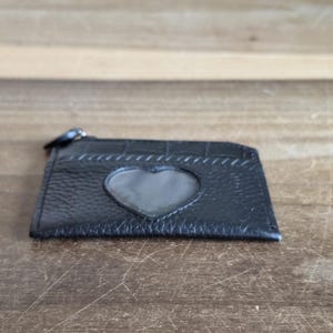 May include: A black leather card holder featuring a textured surface and a heart-shaped clear window on the front. The accessory includes a top zipper closure with a small black pull tab, providing a compact design for storing cards and small items.