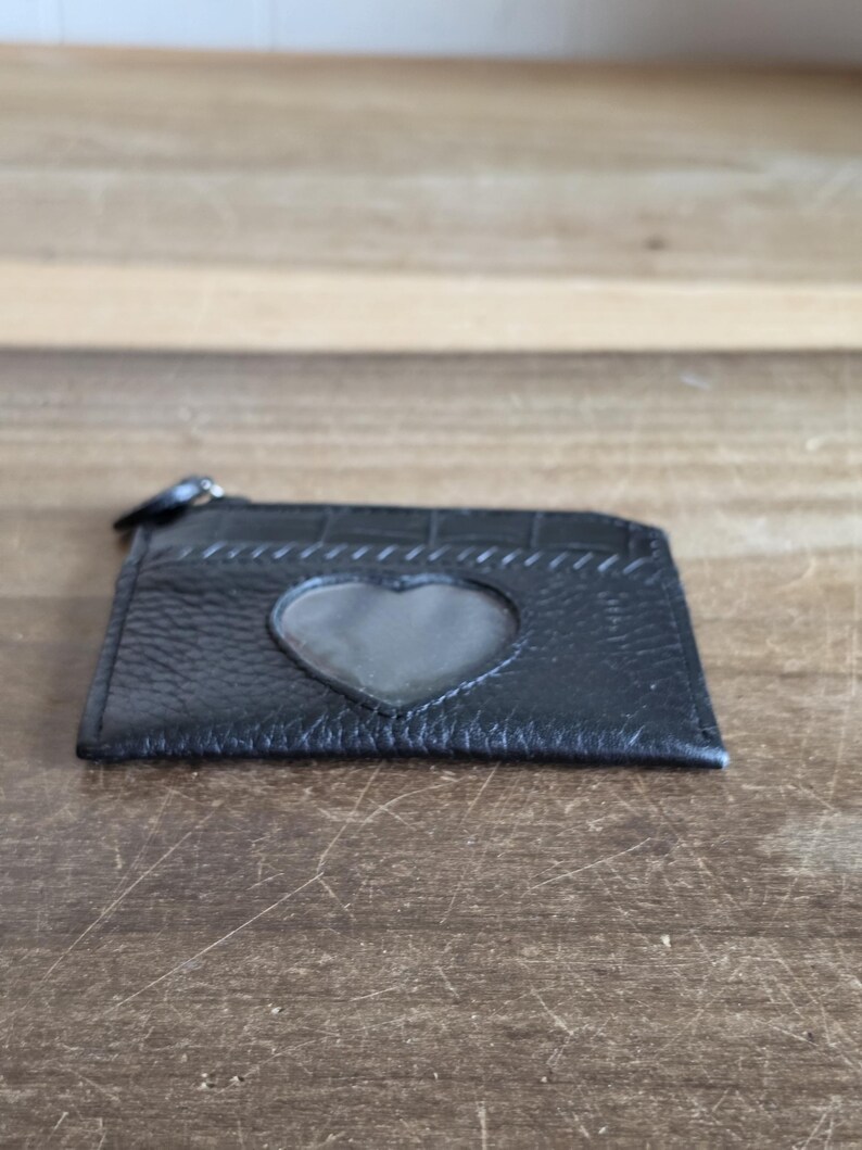 May include: A black leather card holder featuring a textured surface and a heart-shaped clear window on the front. The accessory includes a top zipper closure with a small black pull tab, providing a compact design for storing cards and small items.