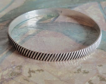 May include: A silver bangle bracelet with a textured, ridged design. The bracelet is simple and elegant.