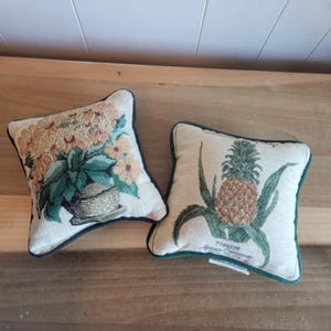 May include: Two small square throw pillows with embroidered floral designs. One pillow features a bouquet of yellow flowers in a green pot, the other features a pineapple with green leaves and the text "Pineapple Agnes  Comstock 1993".