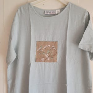 May include: A light blue short-sleeve shirt with a round neckline. The shirt features a square, embroidered patch with a floral design in shades of white and green on a tan background. The label reads "BEDFORD FAIR".